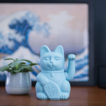 Charger l'image dans la galerie, Figurine Maneki Neko chat porte-bonheur bleu clair posée sur une table en bois, devant une œuvre d'art représentant une vague japonaise et une petite plante verte.
