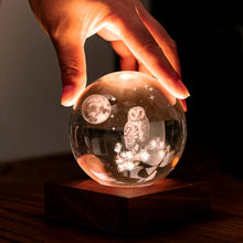 Charger l'image dans la galerie, Main tenant délicatement la veilleuse boule de cristal 3D illuminée révélant une chouette et la lune sur son socle en bois de noyer.
